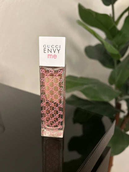 Gucci Envy me perfume bottle on a reflective surface with a plant in the background